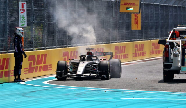 Formula 1 car exiting pit lane with smoke from tires as a crew member watches from the track edge, DHL banners along the barrier.