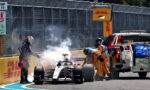 Formula 1 car at the pit wall with smoke and crew members in yellow suits attending, near a DHL barrier and service truck.