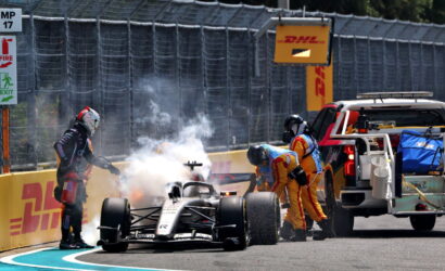 Formula 1 car at the pit wall with smoke and crew members in yellow suits attending, near a DHL barrier and service truck.