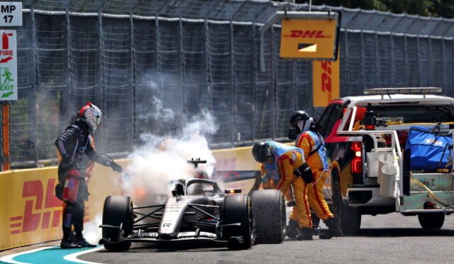 Formula 1 car at the pit wall with smoke and crew members in yellow suits attending, near a DHL barrier and service truck.