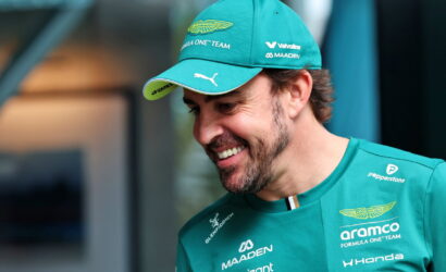 Man in teal Aston Martin cap and Formula One team shirt smiles, looking down to the right, logos visible on clothing and cap.