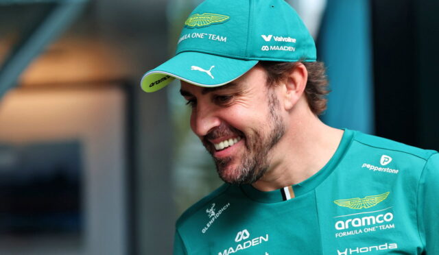 Man in teal Aston Martin cap and Formula One team shirt smiles, looking down to the right, logos visible on clothing and cap.