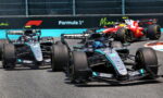 Four Formula 1 cars race on a track; lead car is a Mercedes in teal/black, red Ferrari behind; Formula 1 signage in the background.