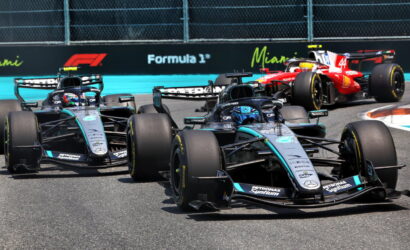Four Formula 1 cars race on a track; lead car is a Mercedes in teal/black, red Ferrari behind; Formula 1 signage in the background.