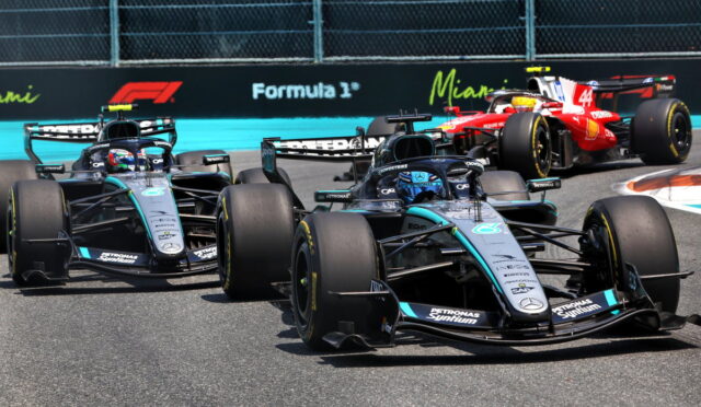 Four Formula 1 cars race on a track; lead car is a Mercedes in teal/black, red Ferrari behind; Formula 1 signage in the background.
