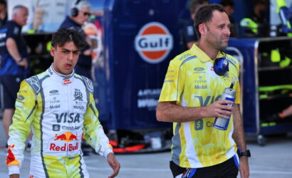 Two Formula 1 drivers in racing suits walk through a pit lane with a Gulf truck in the background, one holding a water bottle and the other looking ahead.
