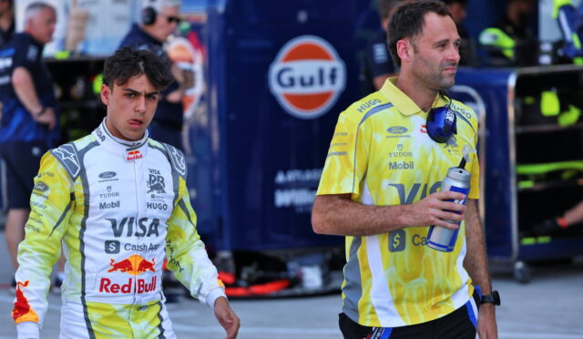 Two Formula 1 drivers in racing suits walk through a pit lane with a Gulf truck in the background, one holding a water bottle and the other looking ahead.