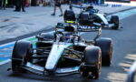 Mercedes-AMG Petronas Formula One car on a race track with a second car behind and pit-club spectators in the background, sponsor logos visible.