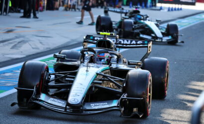 Mercedes-AMG Petronas Formula One car on a race track with a second car behind and pit-club spectators in the background, sponsor logos visible.