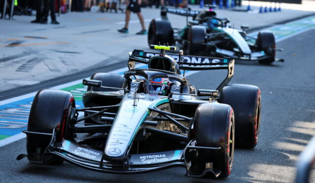 Mercedes-AMG Petronas Formula One car on a race track with a second car behind and pit-club spectators in the background, sponsor logos visible.