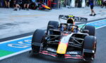 Formula 1 race car on track near pit lane with team members in background and sponsor logos on the car's bodywork (Red Bull/orange nose)