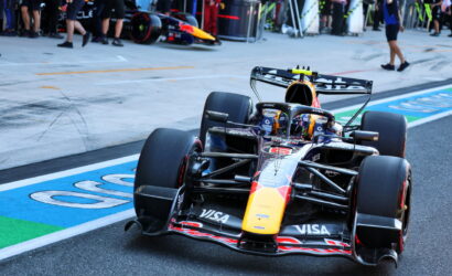 Formula 1 race car on track near pit lane with team members in background and sponsor logos on the car's bodywork (Red Bull/orange nose)