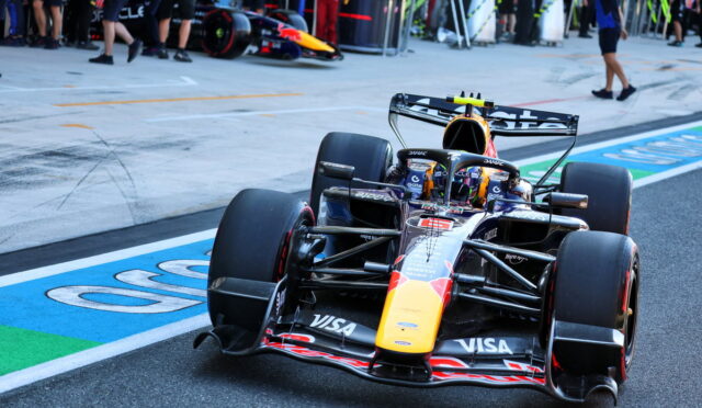 Formula 1 race car on track near pit lane with team members in background and sponsor logos on the car's bodywork (Red Bull/orange nose)