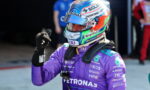 Formula 1 driver in a purple Petronas suit with a colorful helmet, giving a thumbs up.