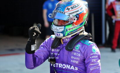Formula 1 driver in a purple Petronas suit with a colorful helmet, giving a thumbs up.