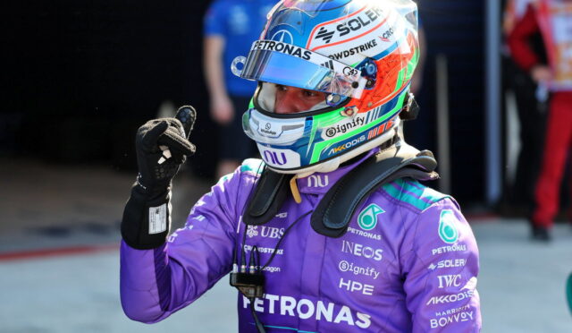 Formula 1 driver in a purple Petronas suit with a colorful helmet, giving a thumbs up.