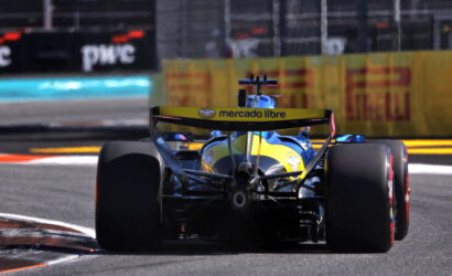 Rear view of a blue-and-yellow Formula racing car on a track, with a Mercado Libre logo on the rear wing and wide tires visible in the foreground.