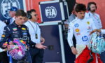 Two Formula 1 drivers stand with helmets in hand near the pit, facing forward with team logos on their suits and crew in the background.