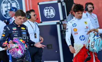 Two Formula 1 drivers stand with helmets in hand near the pit, facing forward with team logos on their suits and crew in the background.
