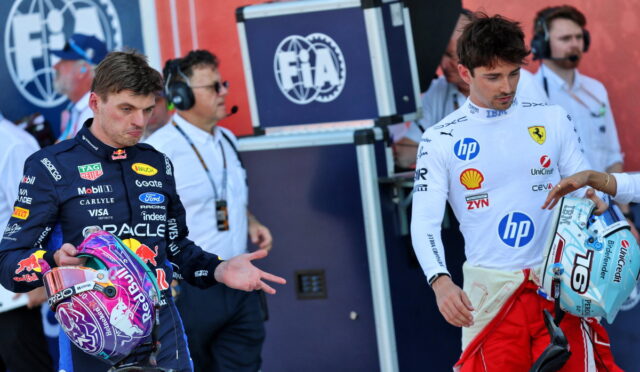 Two Formula 1 drivers stand with helmets in hand near the pit, facing forward with team logos on their suits and crew in the background.