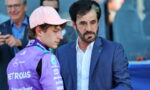 Two men converse at a motorsport event: a driver in a purple racing suit and pink cap beside a man in a blue suit.