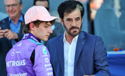 Two men converse at a motorsport event: a driver in a purple racing suit and pink cap beside a man in a blue suit.