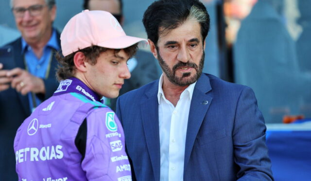 Two men converse at a motorsport event: a driver in a purple racing suit and pink cap beside a man in a blue suit.