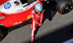 Formula 1 driver in a red racing suit and blue helmet standing beside a red and white race car in the pit lane.