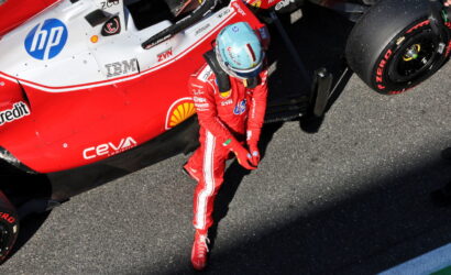 Formula 1 driver in a red racing suit and blue helmet standing beside a red and white race car in the pit lane.