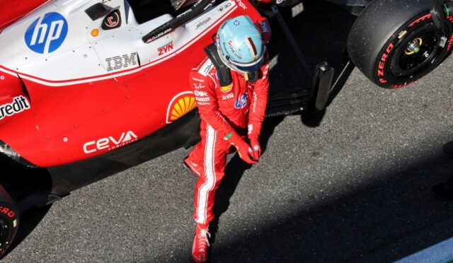 Formula 1 driver in a red racing suit and blue helmet standing beside a red and white race car in the pit lane.