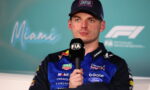 Racing driver in blue Oracle Red Bull Racing suit holding a FIA microphone during a press conference, wearing a cap.