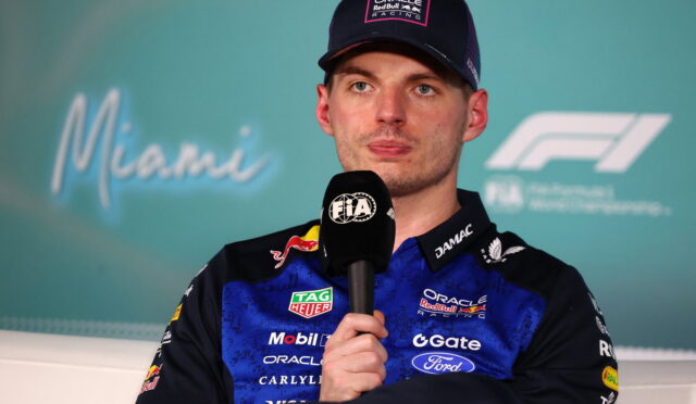 Racing driver in blue Oracle Red Bull Racing suit holding a FIA microphone during a press conference, wearing a cap.
