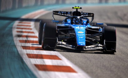 Blue Formula 1 race car speeding around a track, hugging a red-and-white curb at the edge of the road, driver in helmet visible.