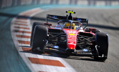 Red Formula 1 race car speeding around a track near red-and-white striped curbs, blurred background suggesting high speed in a corner on a race circuit