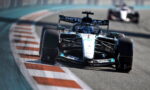 Formula 1 race car on a curving track, foreground with sponsor logos on white car, pursuing a blurred car in the background.
