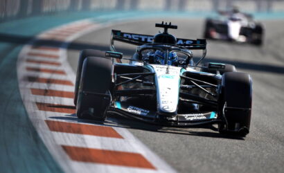 Formula 1 race car on a curving track, foreground with sponsor logos on white car, pursuing a blurred car in the background.