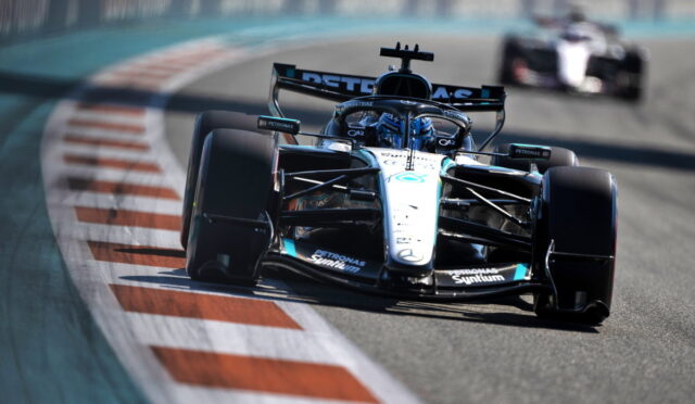 Formula 1 race car on a curving track, foreground with sponsor logos on white car, pursuing a blurred car in the background.