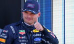 Race car driver in a dark blue suit with sponsor logos, giving a thumbs-up, wearing an Oracle Red Bull Racing cap.