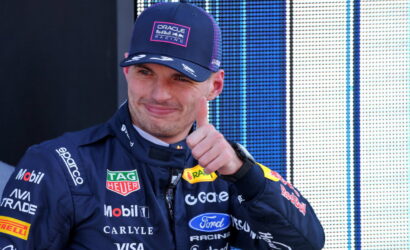 Race car driver in a dark blue suit with sponsor logos, giving a thumbs-up, wearing an Oracle Red Bull Racing cap.