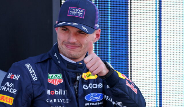 Race car driver in a dark blue suit with sponsor logos, giving a thumbs-up, wearing an Oracle Red Bull Racing cap.