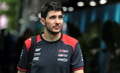 Man with short dark hair and light beard wearing a black and red esports jersey, looking to the side outdoors.