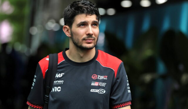 Man with short dark hair and light beard wearing a black and red esports jersey, looking to the side outdoors.