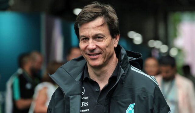 Smiling man in a dark Petronas jacket at an event, with a blurred crowd in the background.