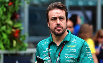 Man with a trimmed beard in a teal polo shirt with sponsor logos, holding a smartphone outdoors and looking thoughtful.