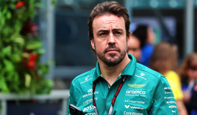 Man with a trimmed beard in a teal polo shirt with sponsor logos, holding a smartphone outdoors and looking thoughtful.