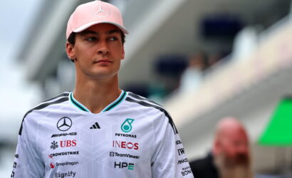 Portrait of a man wearing a pink cap with a Mercedes logo and a white racing polo covered in sponsor logos at a motorsport venue.