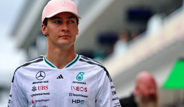 Portrait of a man wearing a pink cap with a Mercedes logo and a white racing polo covered in sponsor logos at a motorsport venue.