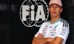 Race car driver with arms crossed in front of a large FIA logo backdrop, wearing a pink Mercedes cap and white sponsor jersey.