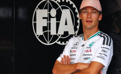 Race car driver with arms crossed in front of a large FIA logo backdrop, wearing a pink Mercedes cap and white sponsor jersey.