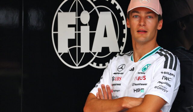 Race car driver with arms crossed in front of a large FIA logo backdrop, wearing a pink Mercedes cap and white sponsor jersey.